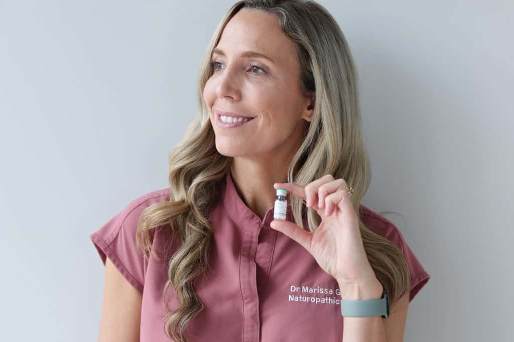 Dr. Marissa Gaucher, Naturopathic Doctor, smiling and holding a small vial of injectable treatment, wearing a clinic uniform against a neutral background.